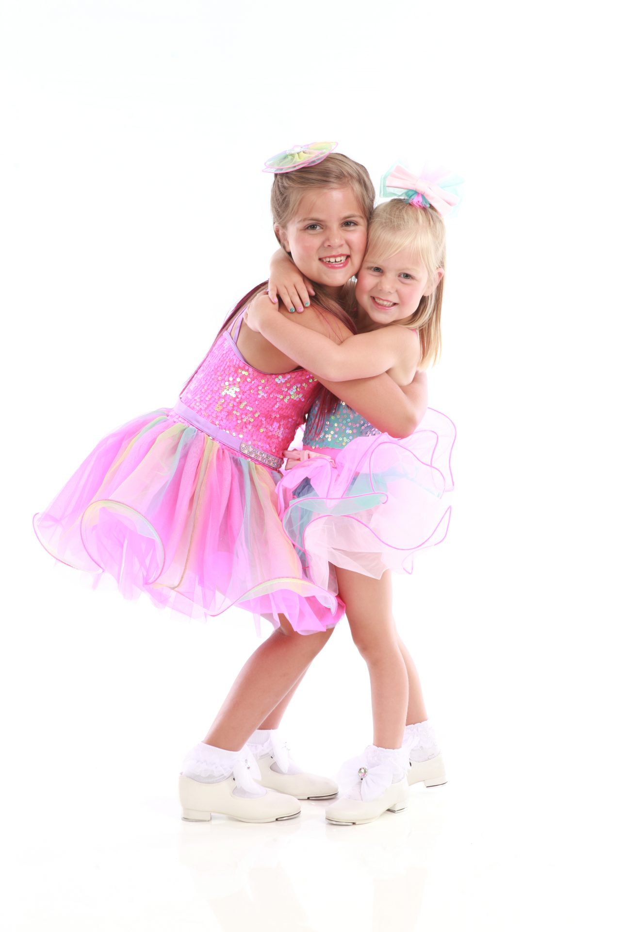 Two young dancers in sequined pink and blue costumes hug each other and pose for dance recital photography in Exulting Images’ Fort Mill SC studio