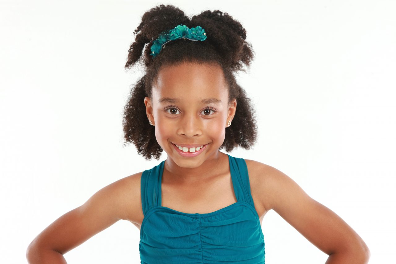 Young female dancer with curly hair pulled up in a half ponytail with a floral clip smiles for dance headshot during dance recital photography in Exulting Images’ Fort Mill SC studio