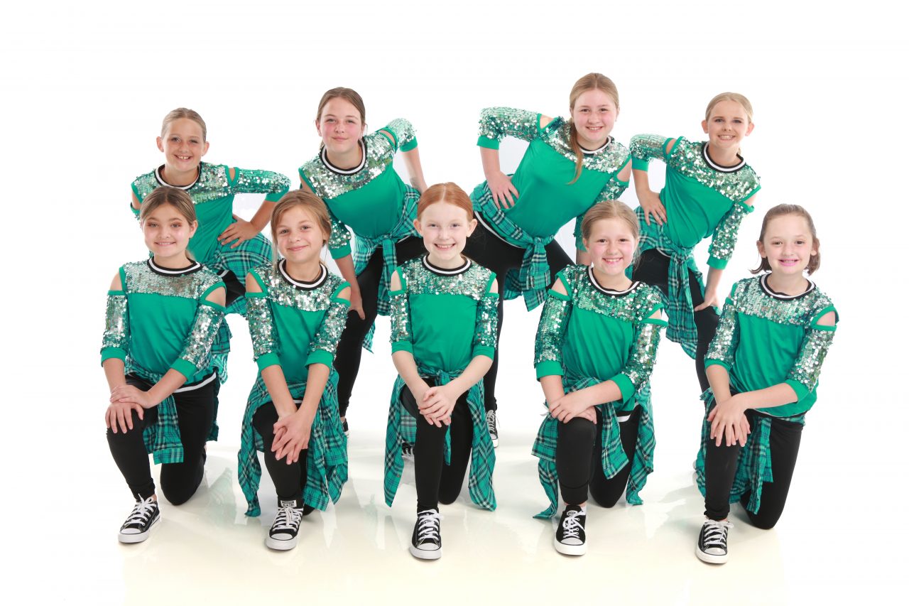 Group of young female dancers wearing sequined green shirts, black bottoms with green flannel shirts tied around their waists, and converse sneakers pose for dance recital photography in Exulting Images’ Fort Mill SC studio