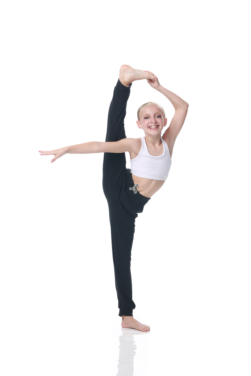 Young female dancer in a white top and black bottoms poses with one leg raised vertically for dance recital photography in Exulting Images’ Fort Mill SC studio
