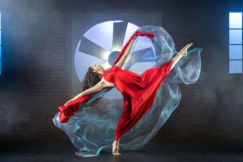 Dancer in red dress poses in front of a large fan.