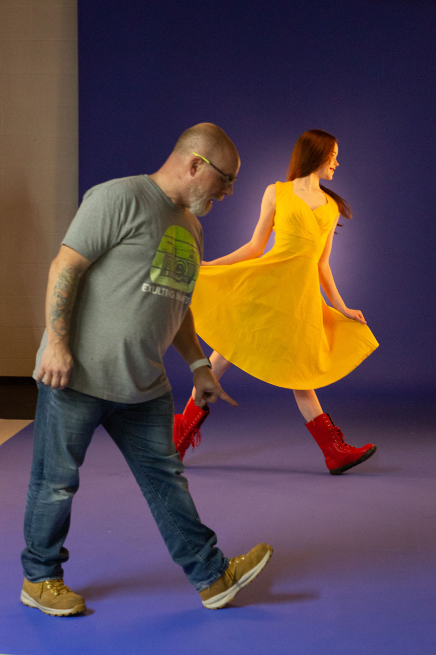 Exulting Images’ President Mike Farella directing a dancer wearing a bright yellow dress and red boots in a pose with toes of front foot lifted for dance photoshoot in Exulting Image’s Fort Mill, SC dance photography studio