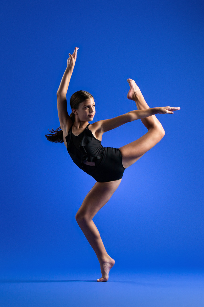 Young female dancer in black leotard with leg in attitude position in front of blue background posing for professional dance photography at Exulting Images’ Fort Mill, SC studio