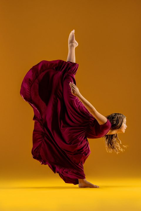 Dance photography portfolio example of female dancer in flowing, voluminous garnet dress poses with one leg lifted steeply behind her for in front of yellow background