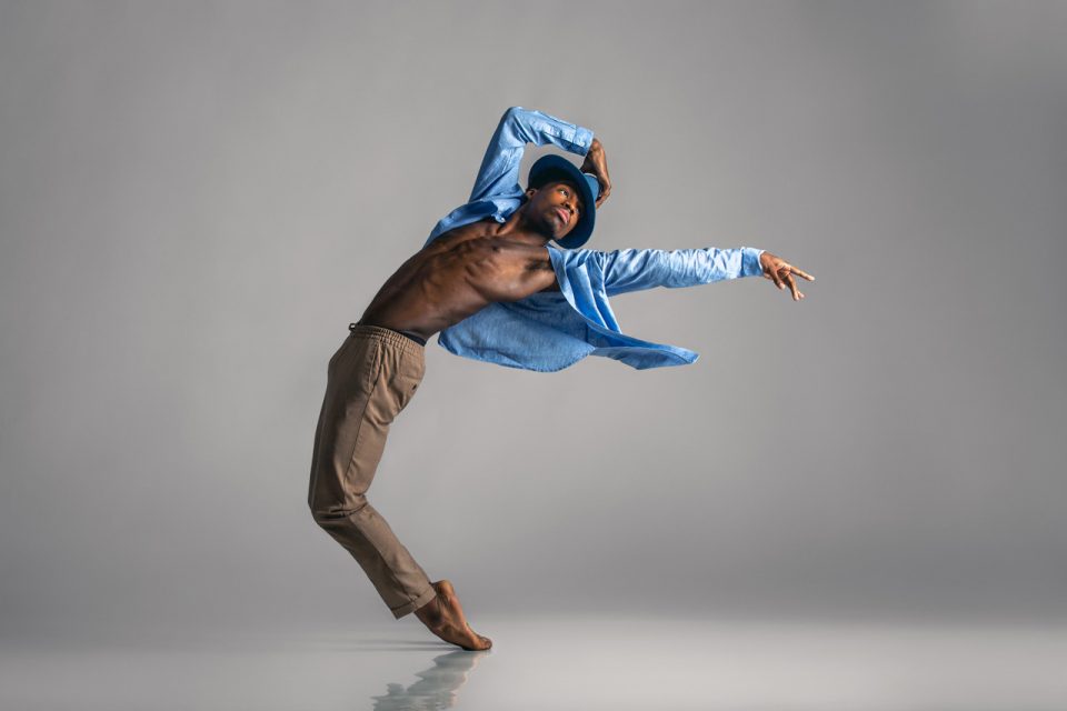 Brown skinned dancer wearing an open blue button up with khaki colored bottoms and blue fedora poses for modern dance action shot in Exulting Images' Fort Mill, SC studio