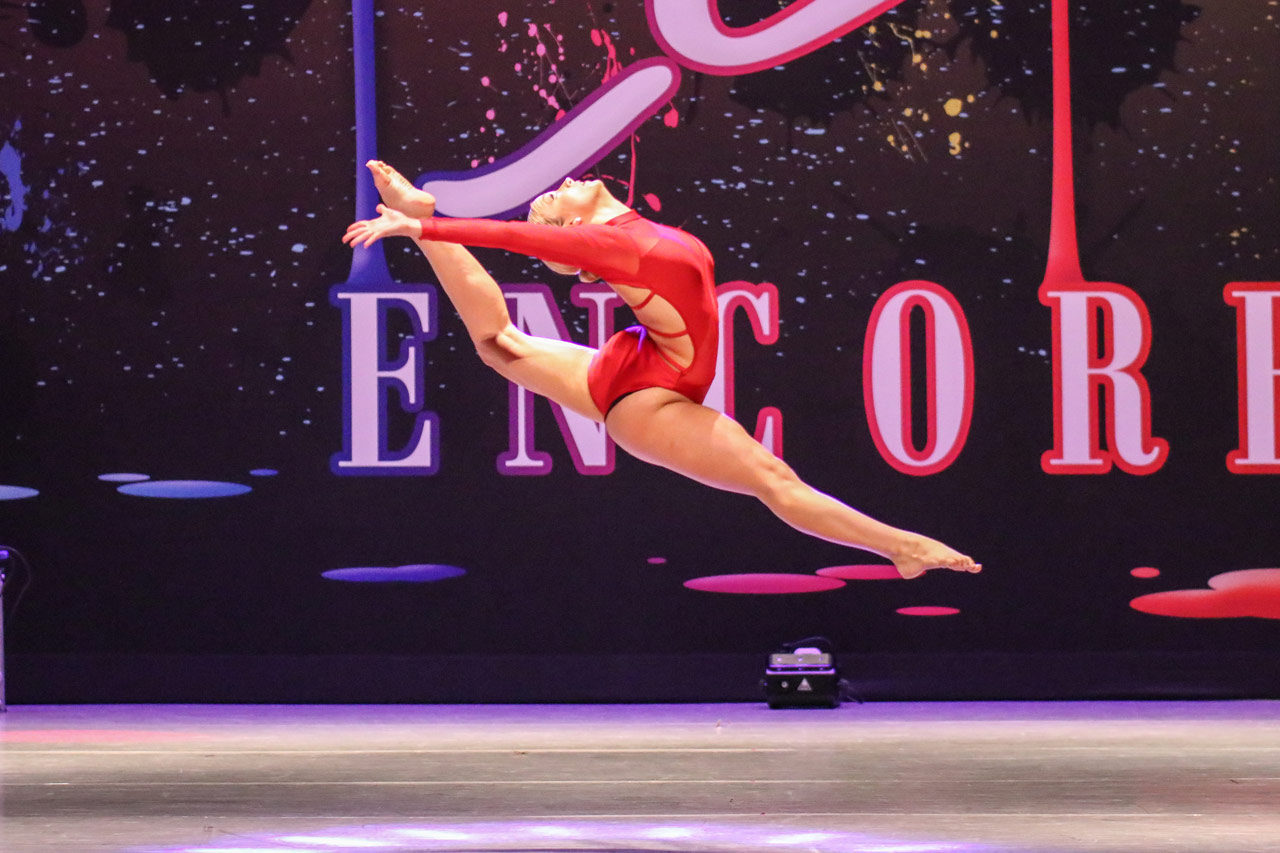 Dance action shot of a dancer wearing a red long sleeved leotard captured by Exulting Images as she is nimbly leaping across the stage during a dance competition