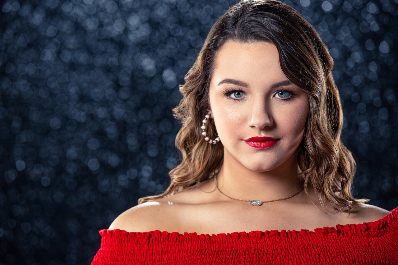 Teenage female with wavy brown hair wearing hoop earrings and sporting bright red lips and a matching off the shoulder blouse stares with captivating hazel eyes for dance headshot in Exulting Images’ Fort Mill, SC studio