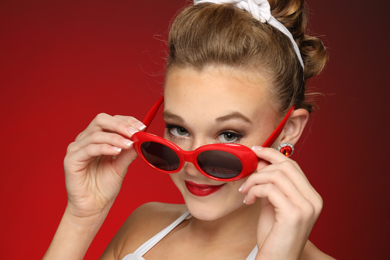 Pre-teen girl with French manicured nails and bright red lips tilts matching bright red sunglasses down to reveal her eyes for headshot taken in Exulting Images’ Fort Mill, SC studio