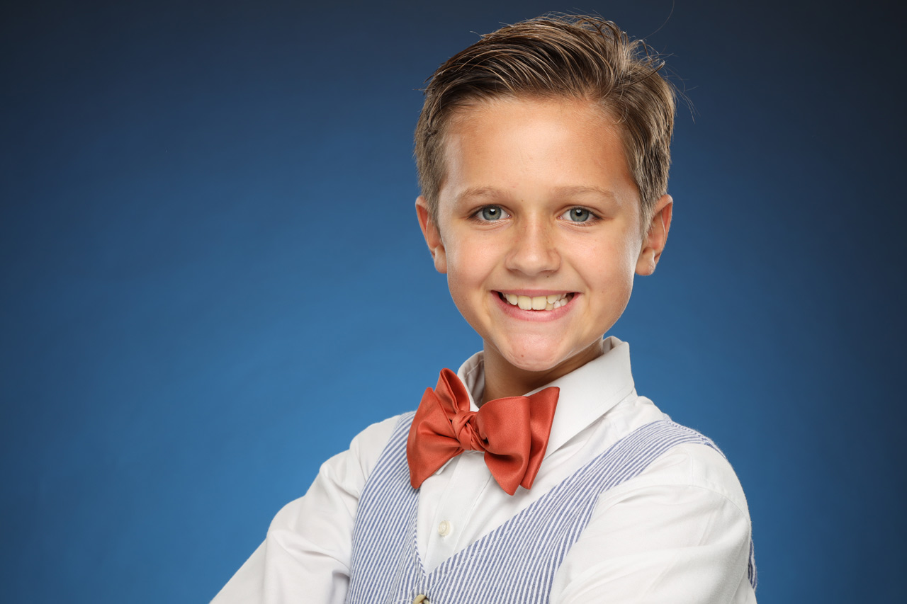 Young boy sporting a bow-time and a seersucker vest smiles at camera in white collared shirt for headshot in Exulting Images’ Fort Mill, SC studio