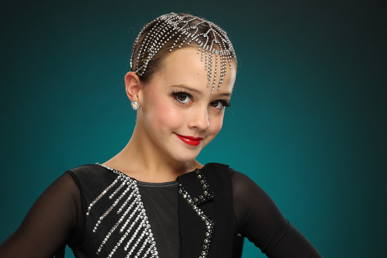 Exulting Images' dancer headshot of young girl wearing rhinestone hair accessory and black dance costume in front of dark teal background