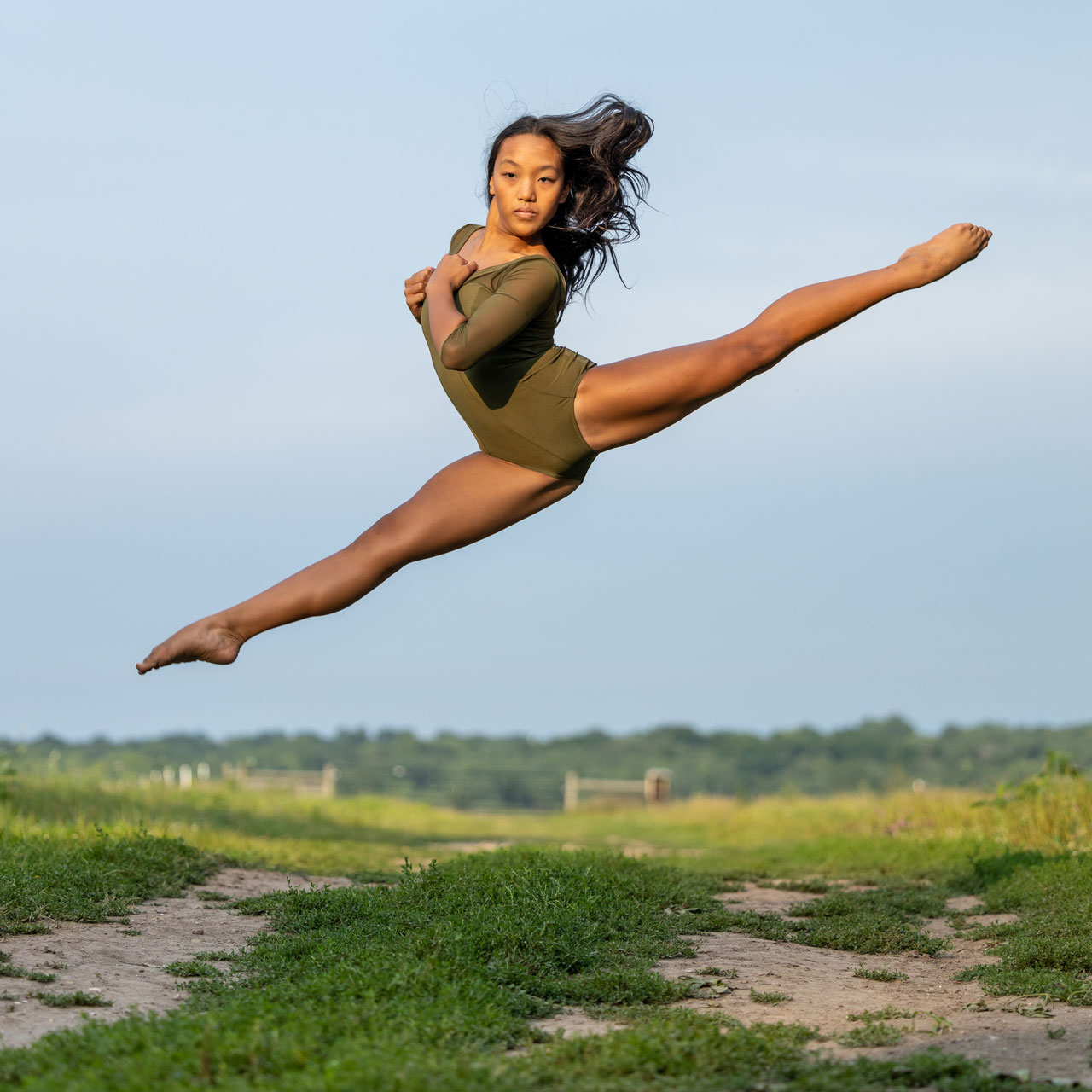 Exulting Images’ outdoor dance action shot of light-skinned dancer in olive green leotard leaping with legs in a split amongst a grassy field