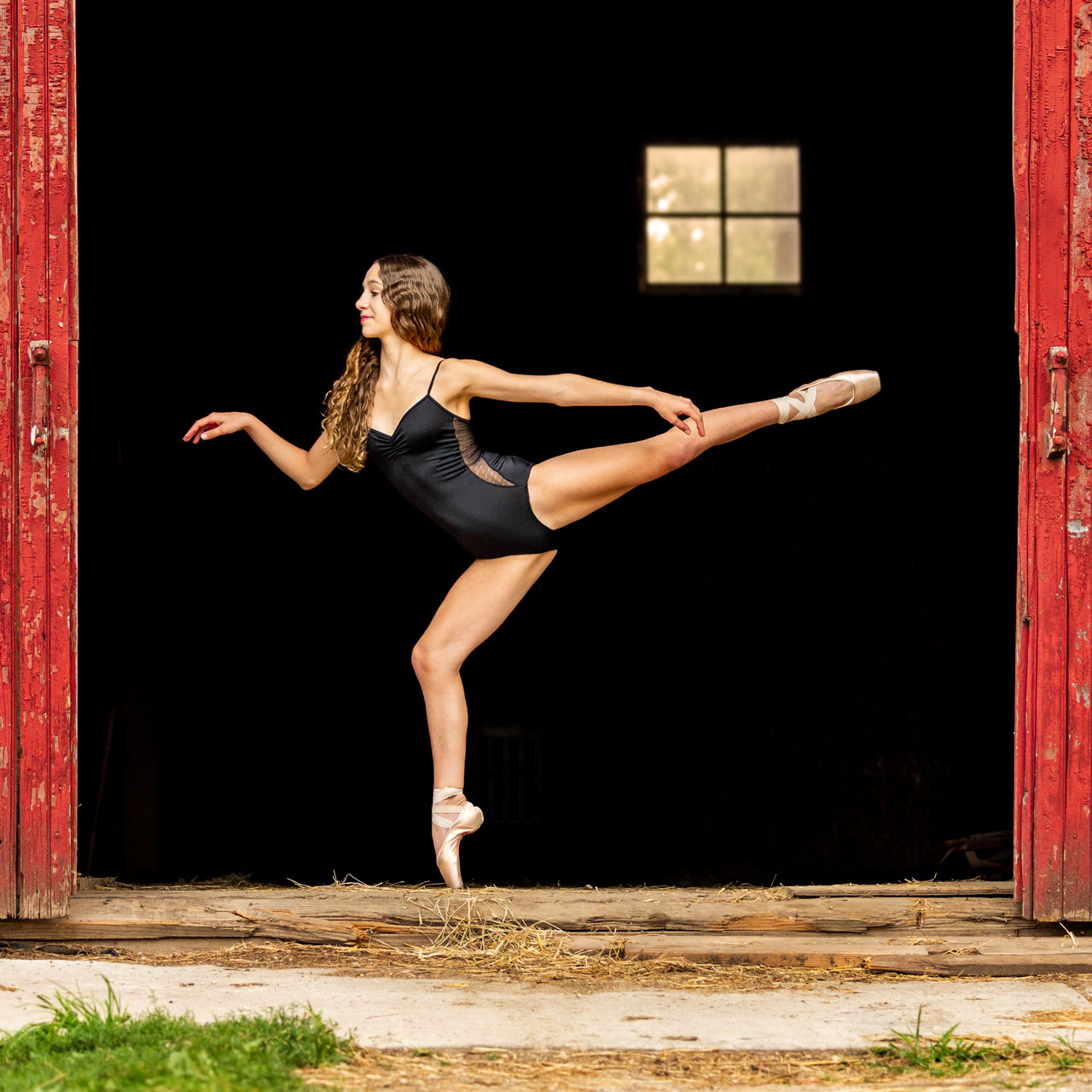 Dancer in black leotard on pointe between two red barn doors in Exulting Images’ dance photoshoot outdoors