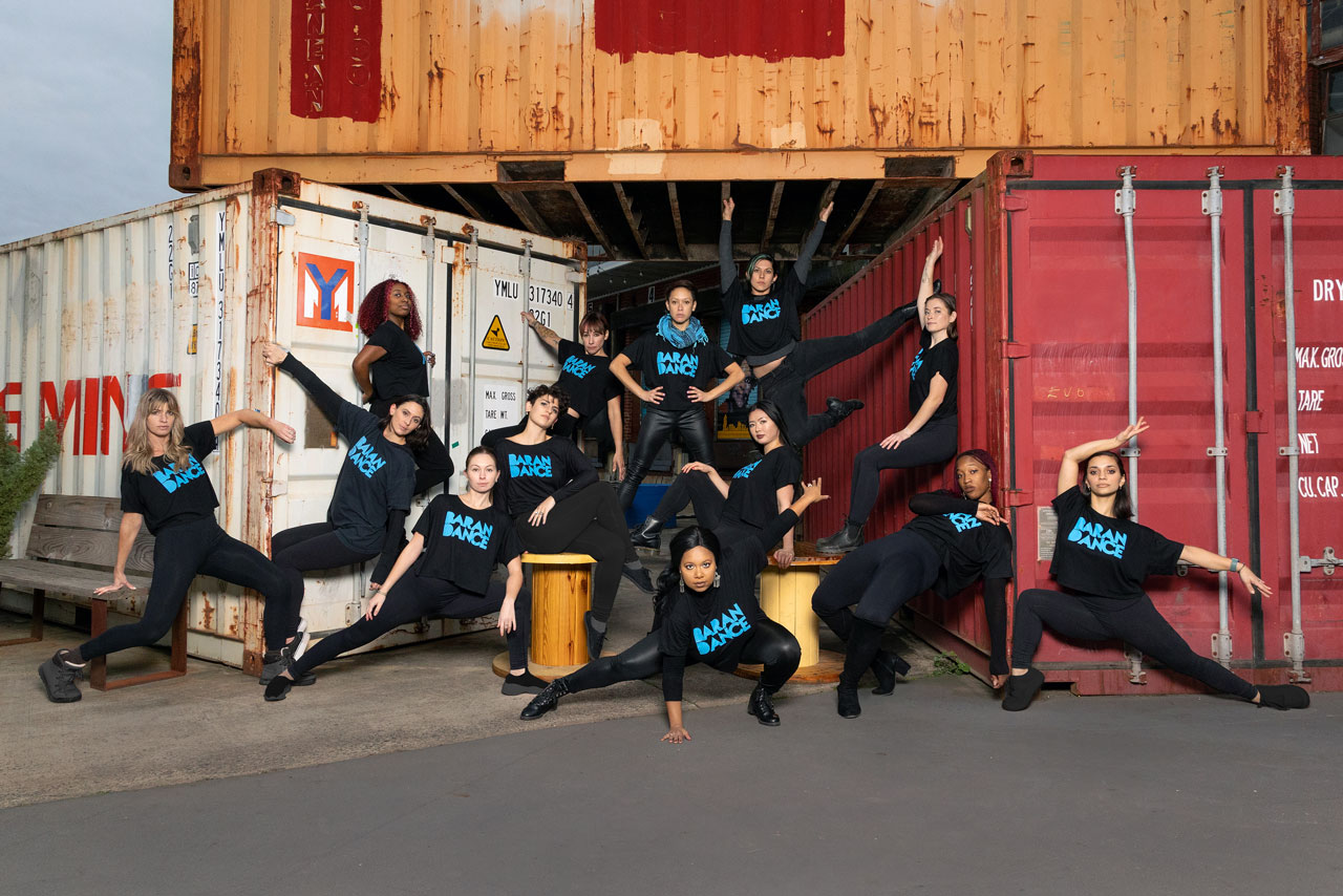 Troupe of contemporary dancers in matching black shirts with the text Baran Dance in bright robin’s blue pose between shipping containers for Exulting Images’ outdoor dance photoshoot