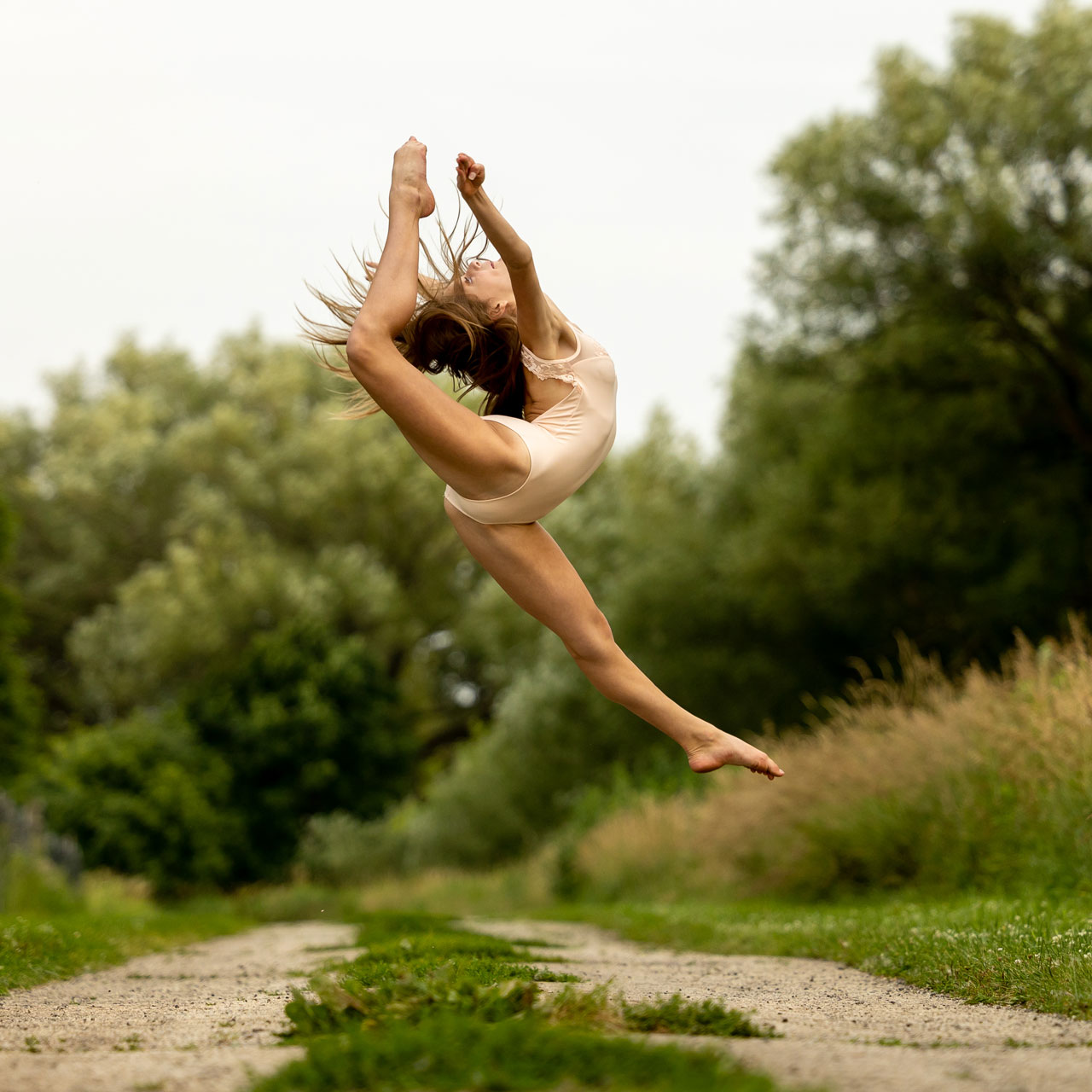 Young dancer in light beige leotard mid leap with leg bent behind her in outdoor dance photoshoot on a dirt road with Exulting Images