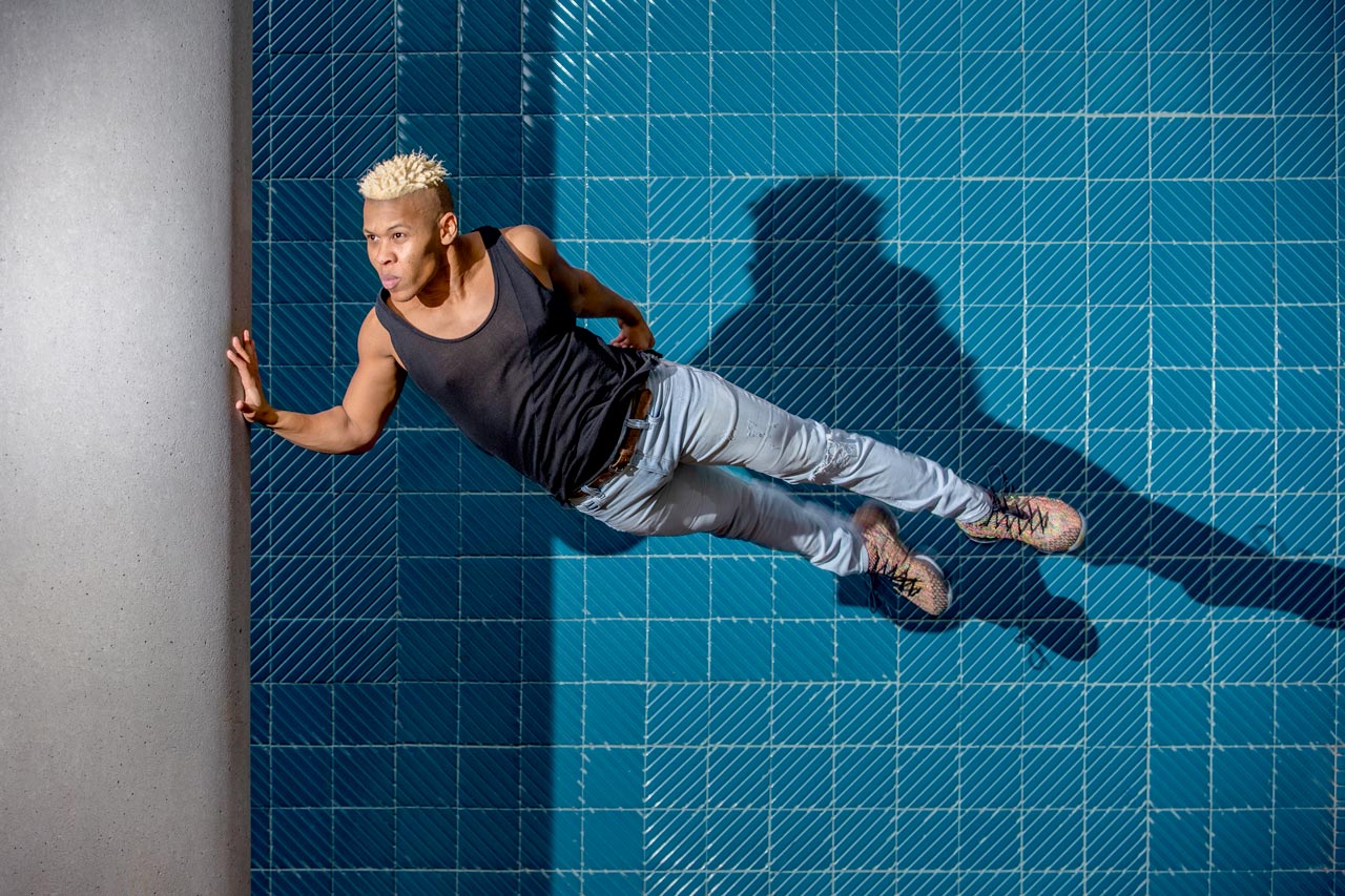 Muscular male dancer with spiked bleach blonde hair poses in front of dark cyan tile with hand gently pressed against a cement pillar during outdoor dance photography session with Exulting Images