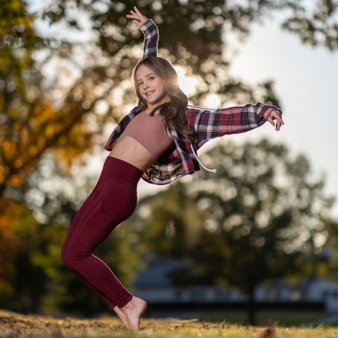 Exulting Images’ outdoor dance photography of young female dancer in maroon leggings and plaid shirt posing on tip toe in front of blurry trees