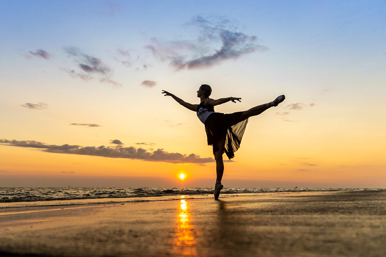 Professional outdoor ballet photo of female in arabesque on a beach as the sun sets behind the ocean