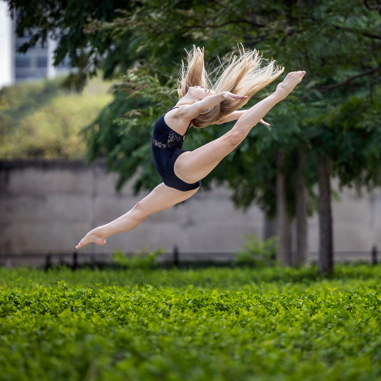 During an Exulting Images’ dance photoshoot outdoors a blonde dancer wearing a navy blue leotard leaps across a blurred lush green background of bushes and trees