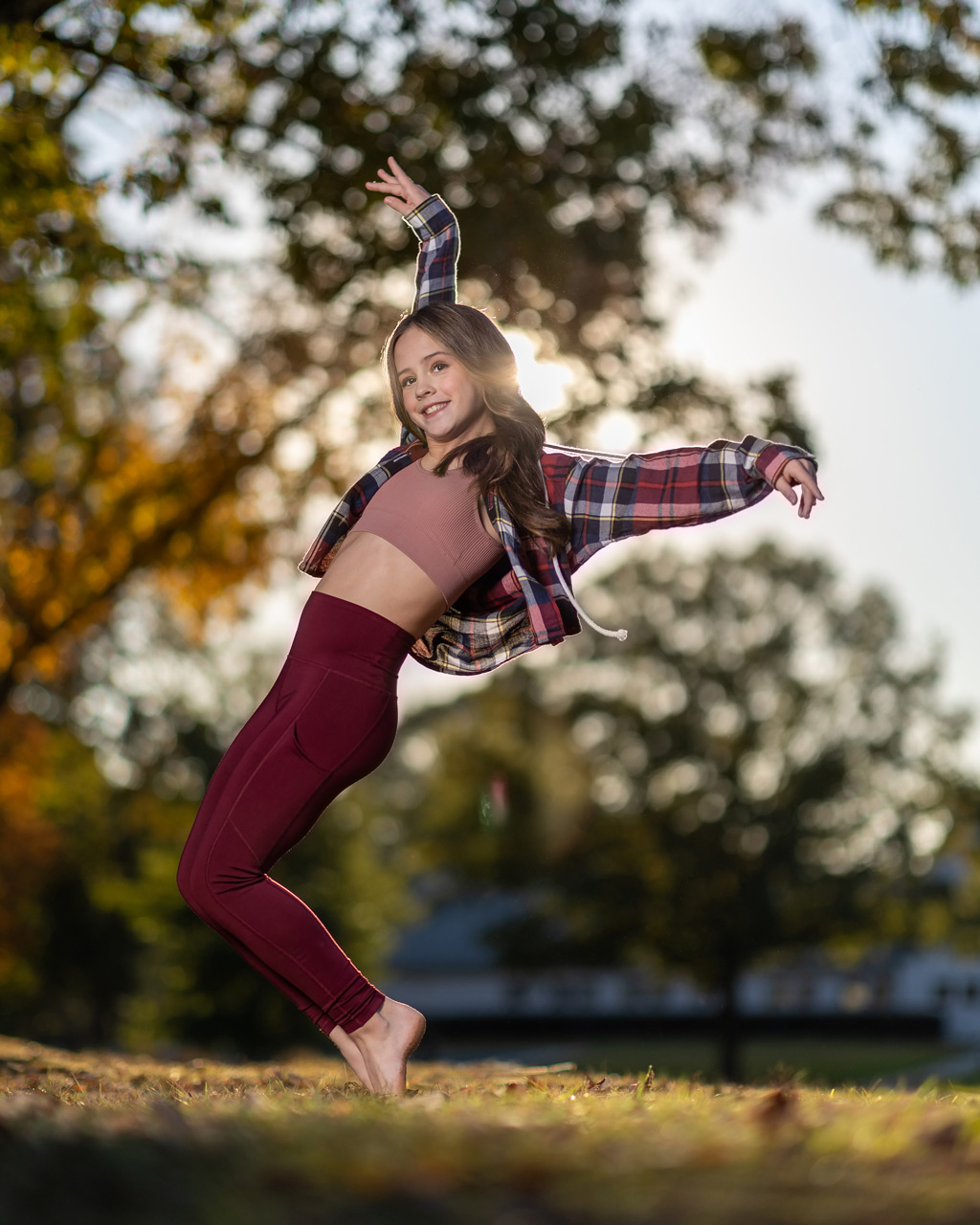 Exulting Images’ outdoor dance photography of young female dancer in maroon leggings and plaid shirt posing on tip toe in front of blurry trees