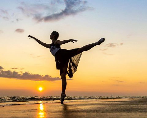 Professional outdoor ballet photo of female in arabesque on a beach as the sun sets behind the ocean