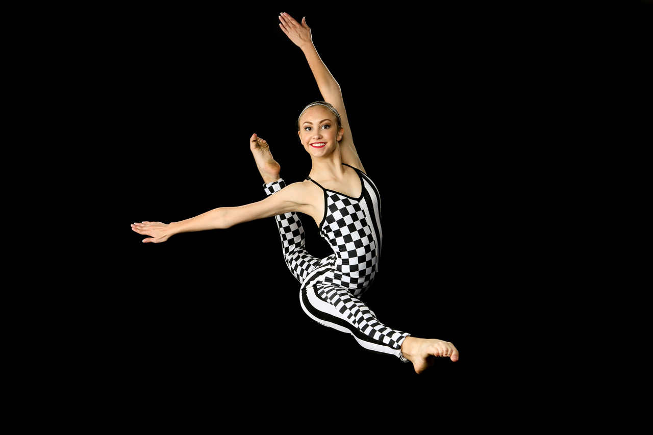 Dance action shot of young female dancer in black and white alternating checkered and striped costume leaping for Exulting Images’ dance recital photography