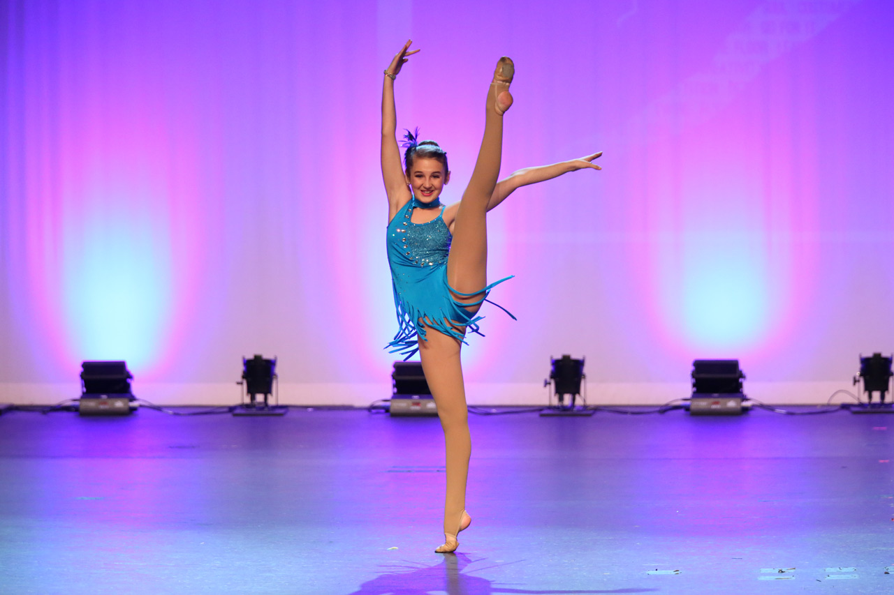 Solo female dancer in aquamarine fringed costume with leg lifted vertically on stage during dance recital photographed by Exulting Images