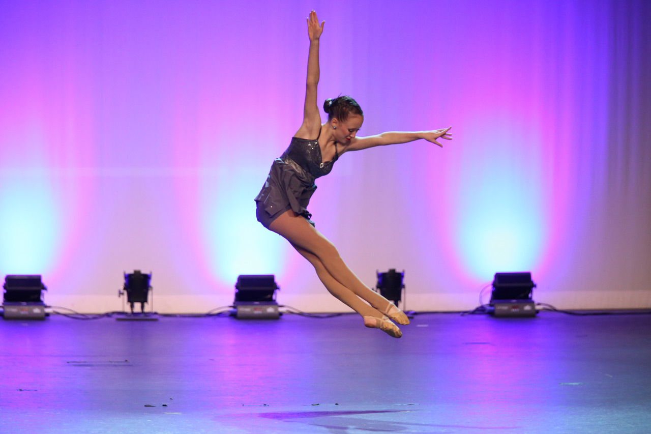 Exulting Images’ dance action shot of young female dancer in a jump with arms forming a ninety degree angle, legs crossed, and toes pointed on stage during a dance recital