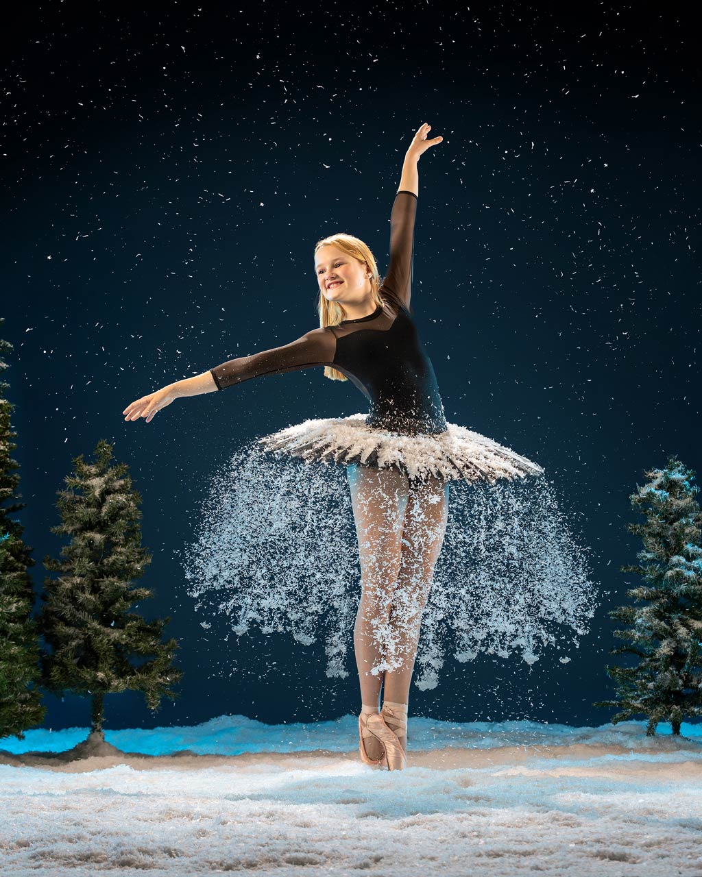 Exulting Images ballet photography of dancer in black tutu dusted with snow next to small evergreens trees