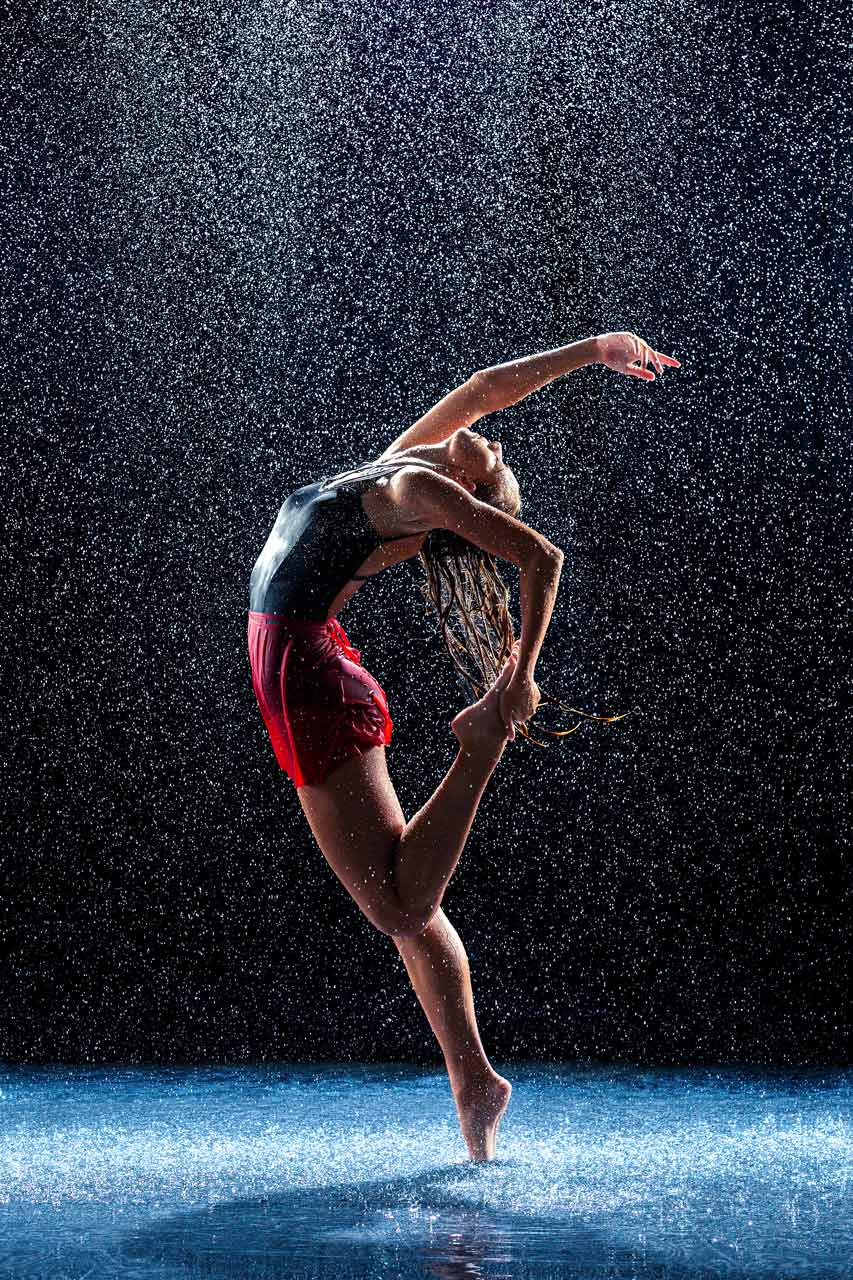 Female dancer poses on tip toe with her back arched and one leg grasped behind in rain room photoshoot at Exulting Images Fort Mill, SC studio