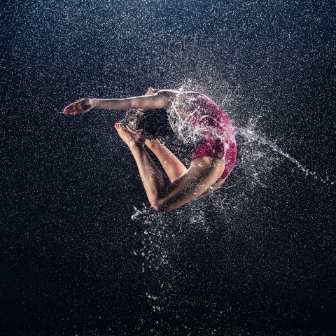 Exulting Images rain dance action shot of dancer in dark pink leotard performing high jump with backbend surrounded by droplets of falling water