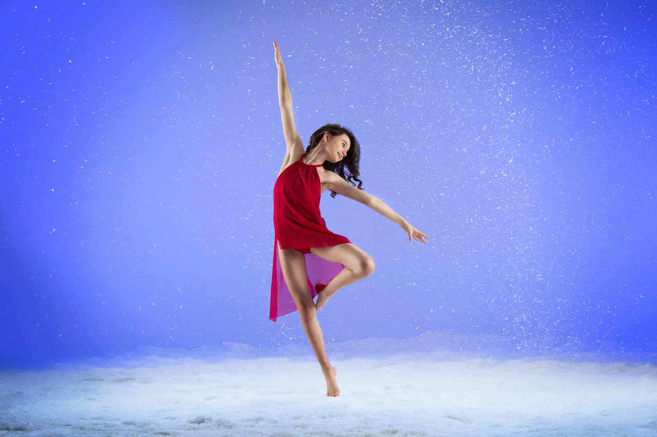 Young brunette dancer in red flowing costume with one leg in passé dances in snow session at Exulting Images Fort Mill, SC professional dance photography studio