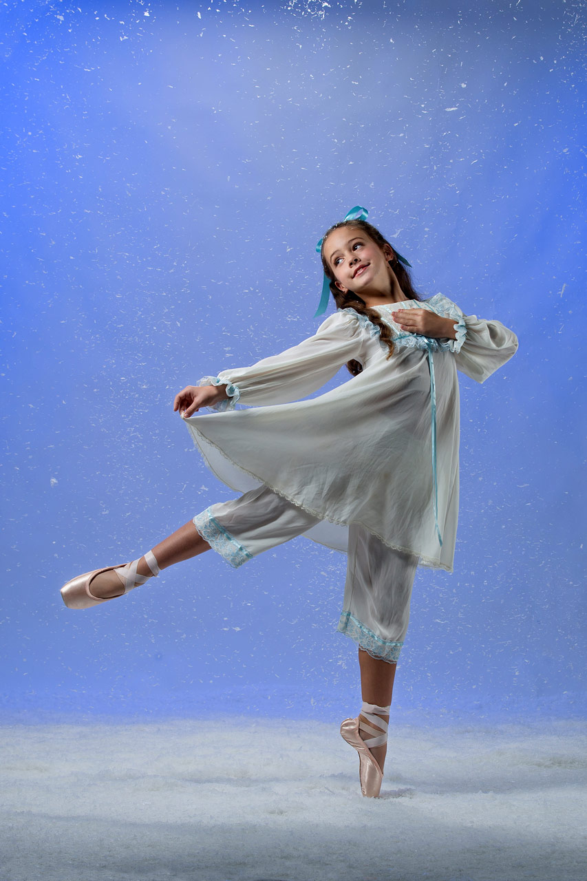 Young ballerina on pointe in white frock with blue silk accents dances in snow session at Exulting Images Fort Mill, SC professional dance photography studio