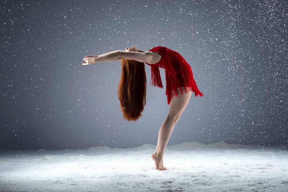 Young auburn haired dancer in red tasseled costume on tip toe reaches backward in a standing back bend n snow session at Exulting Images Fort Mill, SC professional dance photography studio